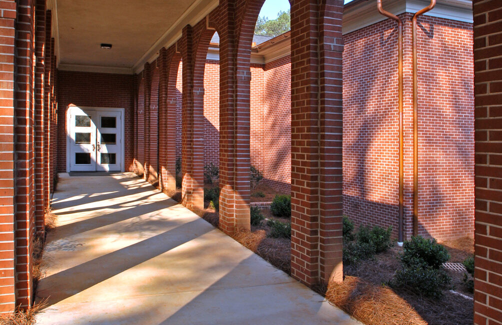 A covered walkway with brick arches and concrete floor leads to a glass door. Sunlight creates shadows; shrubs line the building.