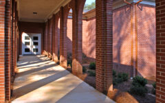A covered walkway with brick arches and concrete floor leads to a glass door. Sunlight creates shadows; shrubs line the building.