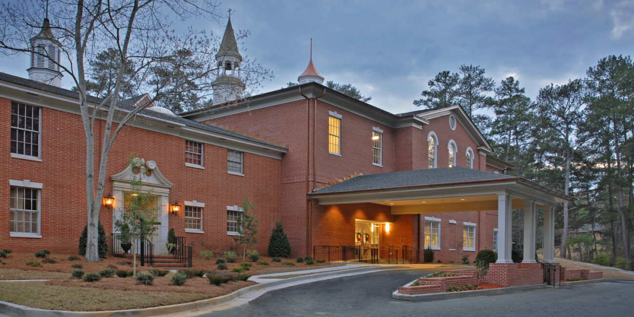 A grand brick building with white entrance columns, tall windows, cupolas on the roof, and trees beneath a cloudy sky.