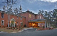 A grand brick building with white entrance columns, tall windows, cupolas on the roof, and trees beneath a cloudy sky.