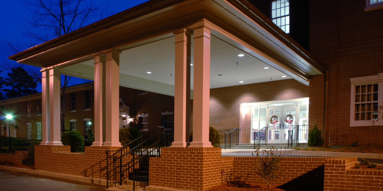 Brick building with a white-columned entrance, outdoor lights at dusk, wreaths on glass doors, and bushes along the walkway.