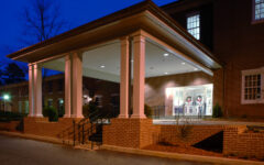 Brick building with a white-columned entrance, outdoor lights at dusk, wreaths on glass doors, and bushes along the walkway.