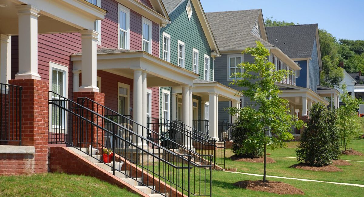 Colorful modern townhouses with front porches, brick steps, and railings on a grassy lawn with young trees and a sunny sidewalk.