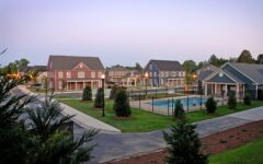 Suburban neighborhood at dusk, colorful houses, trimmed lawns, small trees, and a fenced pool. Winding paths and driveways.