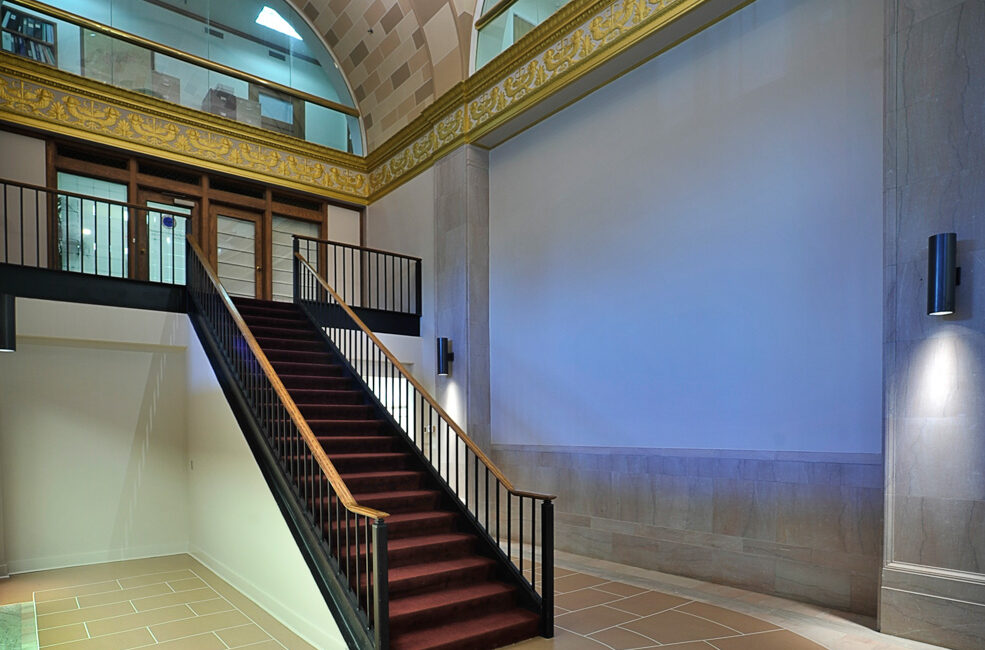 A grand staircase with dark railings and red carpet leads to a mezzanine in a hall with arched ceilings and gold-trimmed cornice.