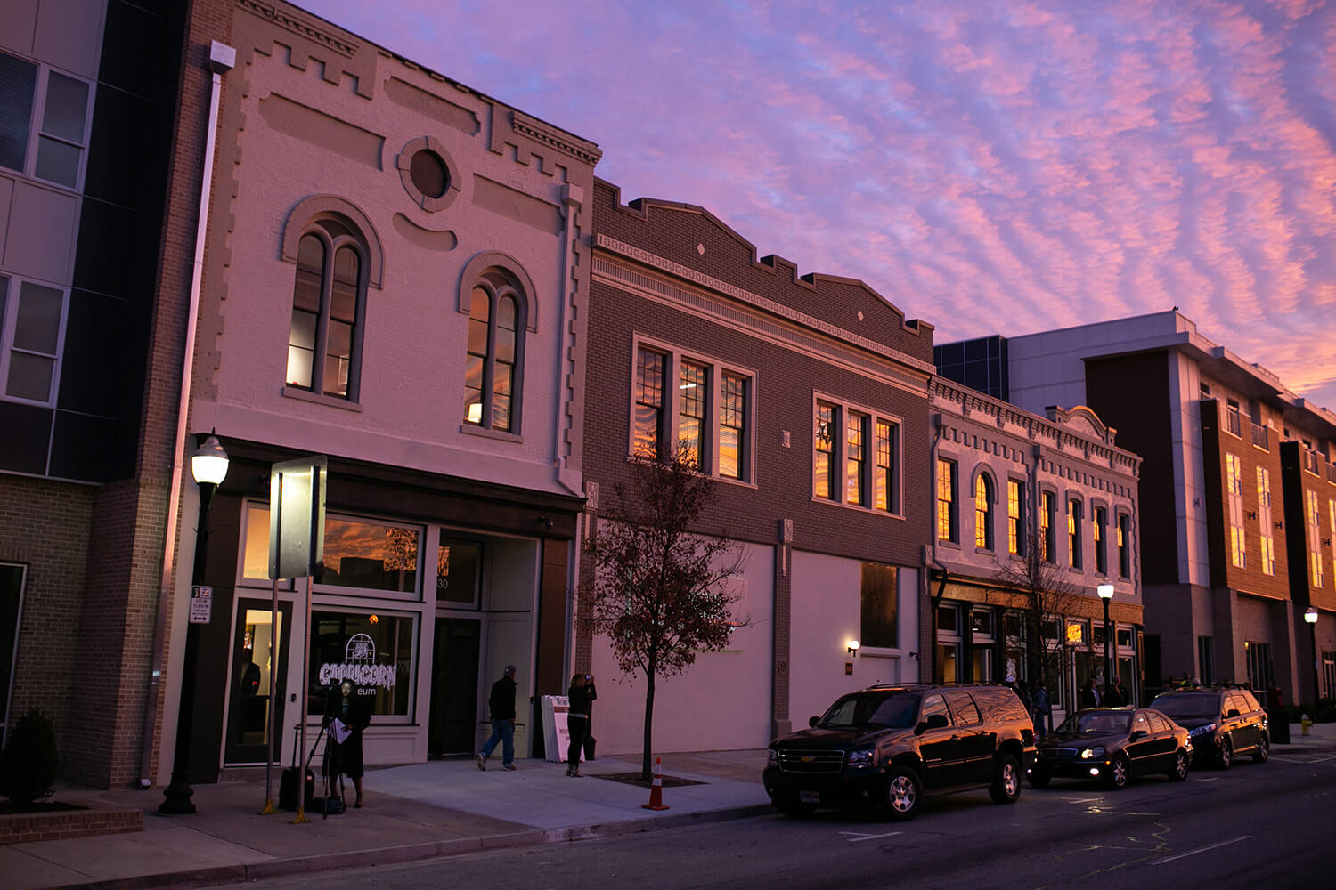 Historic and modern buildings line a sunset street with purple-pink clouds; people walk on the sidewalk, cars park at the curb.