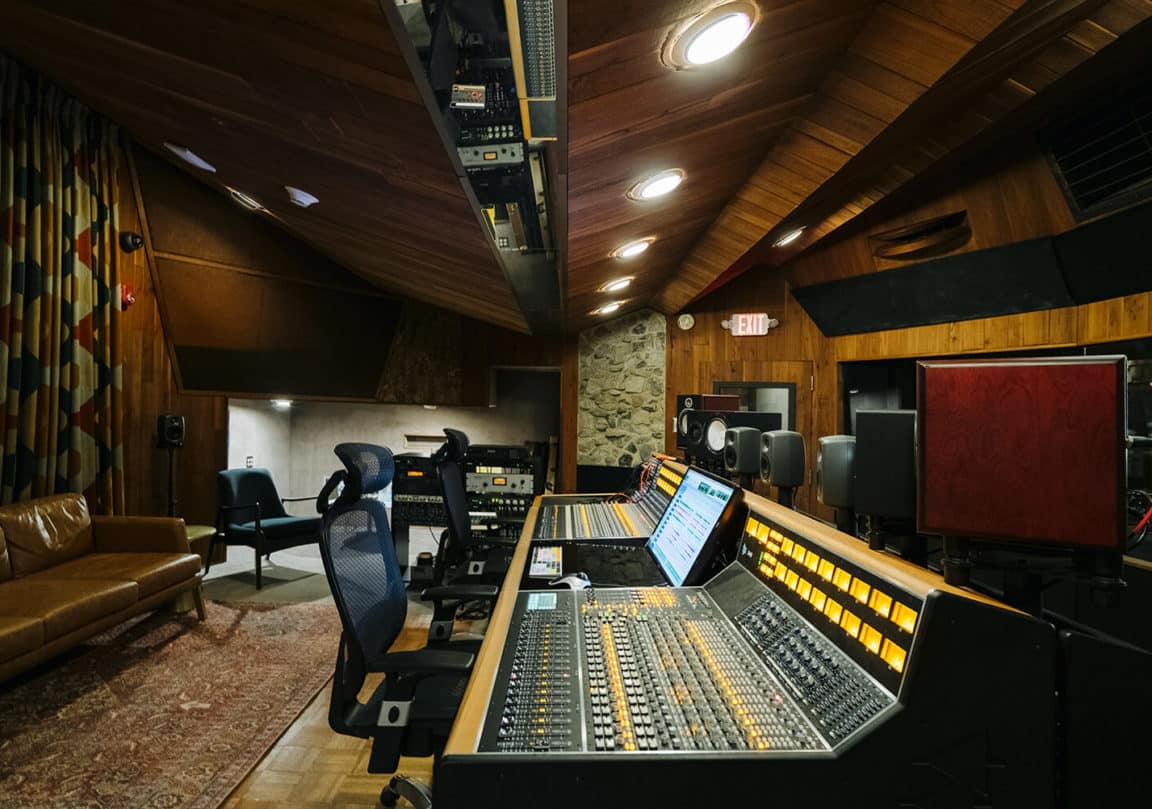 A recording studio featuring large mixing consoles, chairs, audio gear, wood panels, a patterned curtain, and a brown couch.