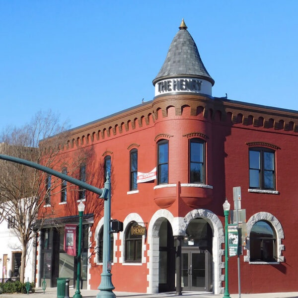 A red brick building with a turret and arched windows, featuring a sign reading THE HENRY near the roof. Trees and streetlights line the sidewalk under a clear blue sky.