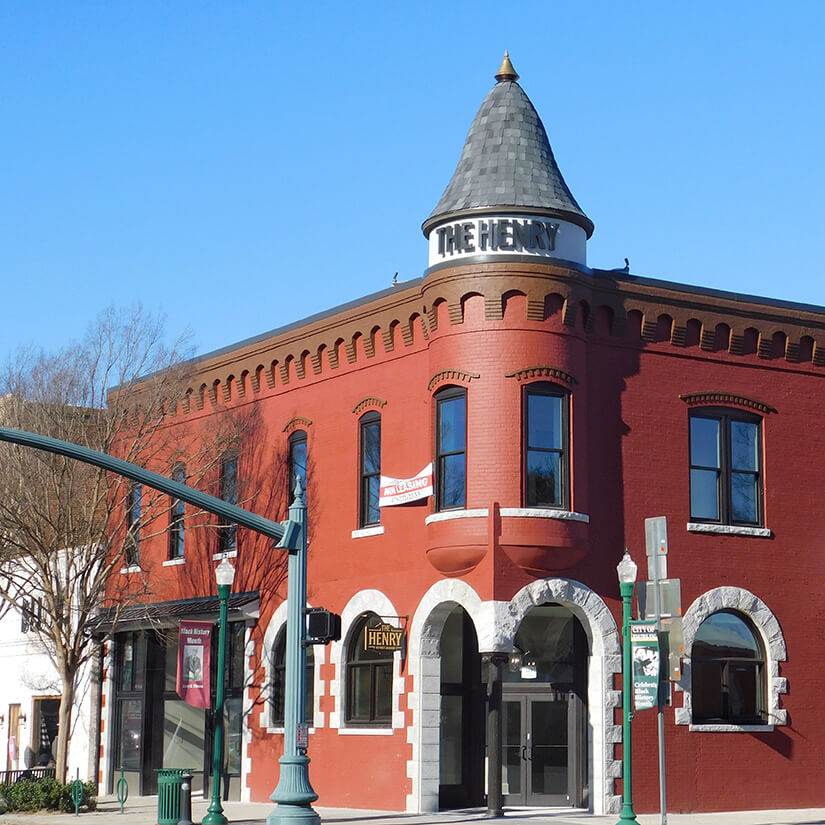 A red brick building with a turret and arched windows, featuring a sign reading THE HENRY near the roof. Trees and streetlights line the sidewalk under a clear blue sky.