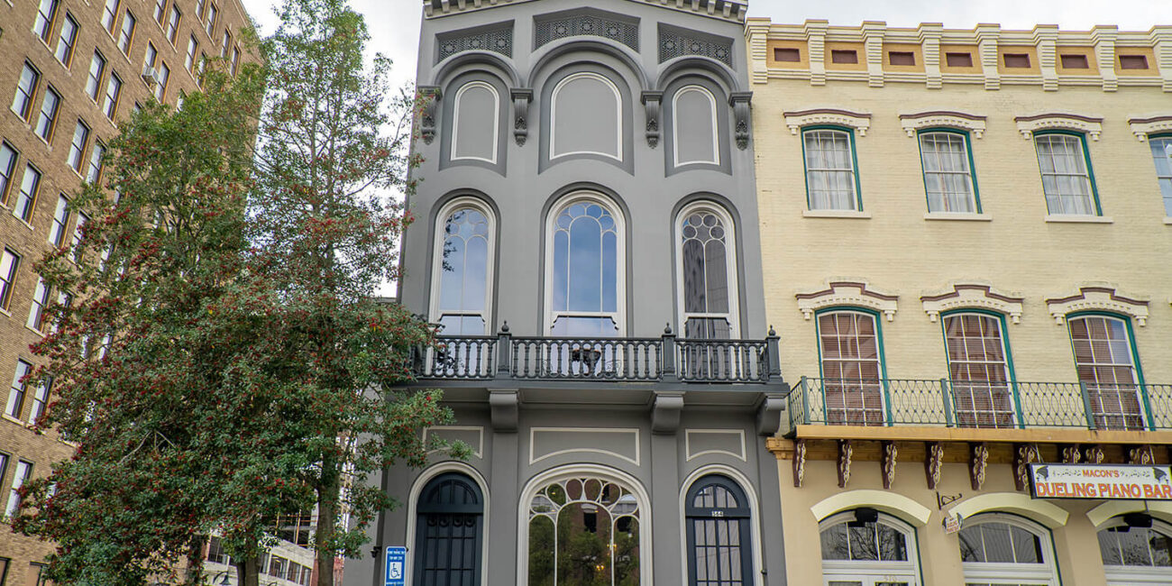 Tall, narrow gray building with arched windows stands between a beige historic building and tree; ornate trim and black balcony above door.