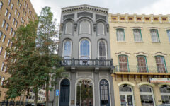 Tall, narrow gray building with arched windows stands between a beige historic building and tree; ornate trim and black balcony above door.