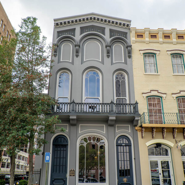 Tall, narrow gray building with arched windows stands between a beige historic building and tree; ornate trim and black balcony above door.