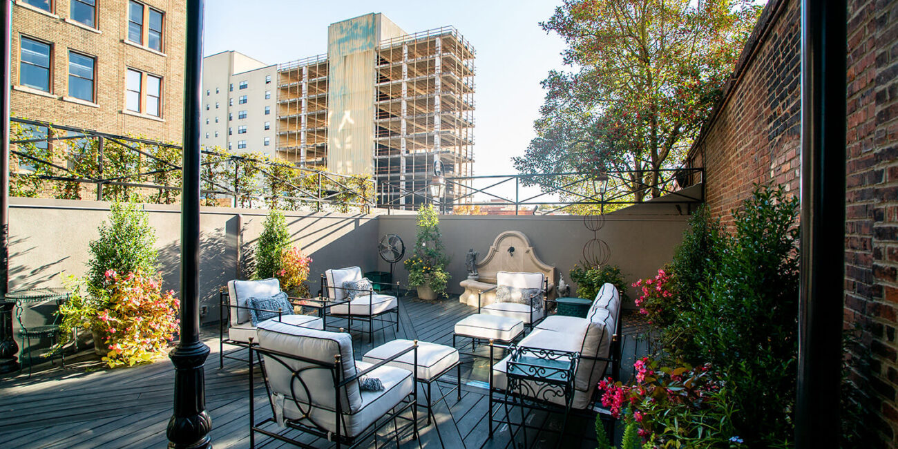 A stylish patio with cushioned metal chairs, glass tables, plants, flowers, a wall fountain, and city views under a clear sky.