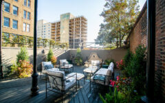 A stylish patio with cushioned metal chairs, glass tables, plants, flowers, a wall fountain, and city views under a clear sky.