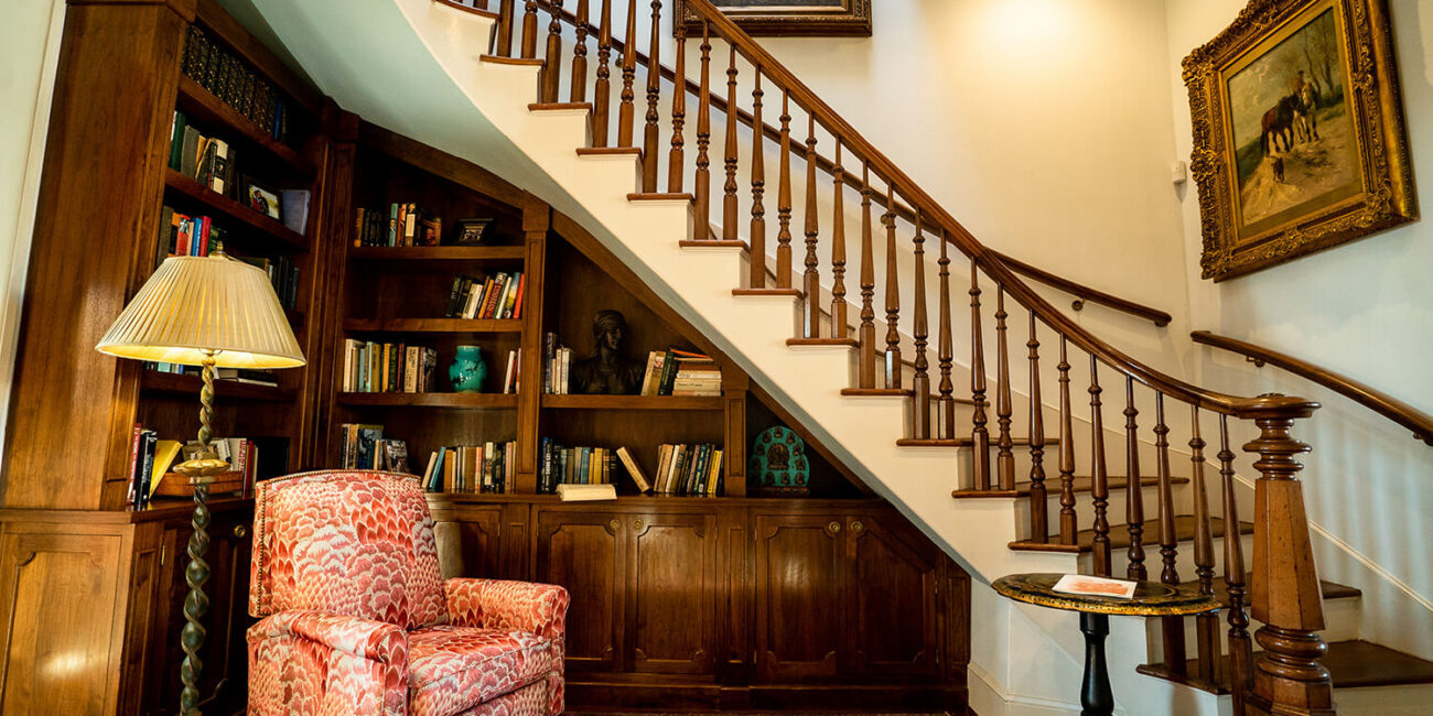 Cozy reading nook under wooden stairs with built-in bookshelves, red armchair, lamp, side table, art on cream walls, and rug.