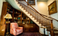 Cozy reading nook under wooden stairs with built-in bookshelves, red armchair, lamp, side table, art on cream walls, and rug.