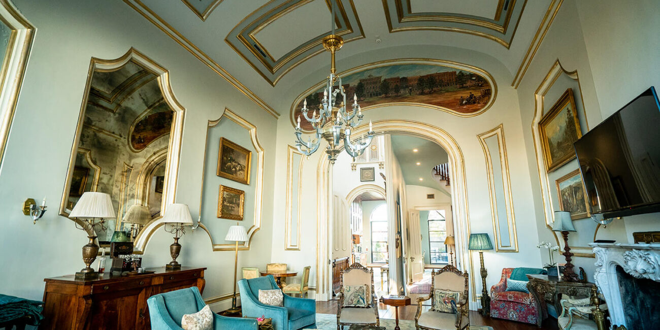 Elegant living room with arched ceilings, gold accents, antique furniture, chandeliers, mirrors, large windows, and blue sofas.