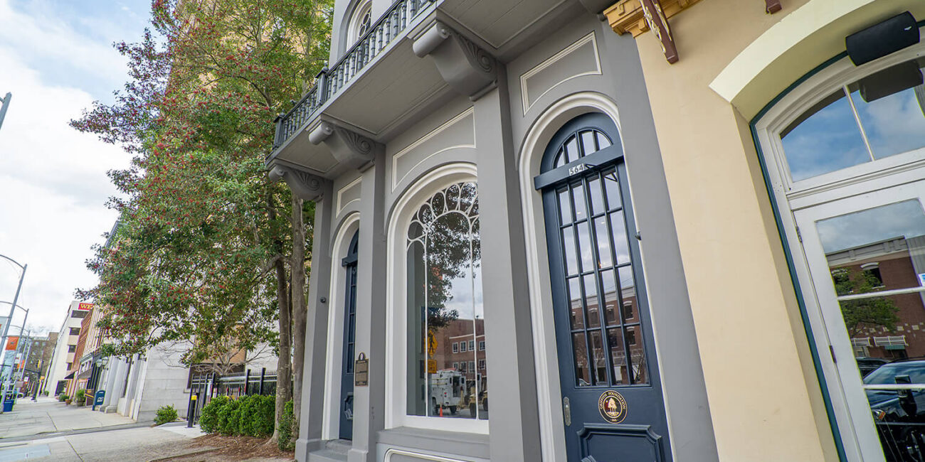A historic gray building with arched windows and a black door on a city sidewalk, a tree to the left, buildings reflected in glass.