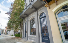 A historic gray building with arched windows and a black door on a city sidewalk, a tree to the left, buildings reflected in glass.