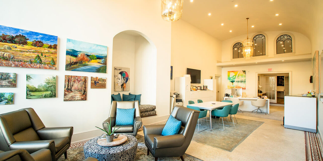 Bright, modern lounge with armchairs, round coffee table, landscape paintings, teal dining chairs, and large hanging lights.