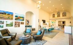 Bright, modern lounge with armchairs, round coffee table, landscape paintings, teal dining chairs, and large hanging lights.