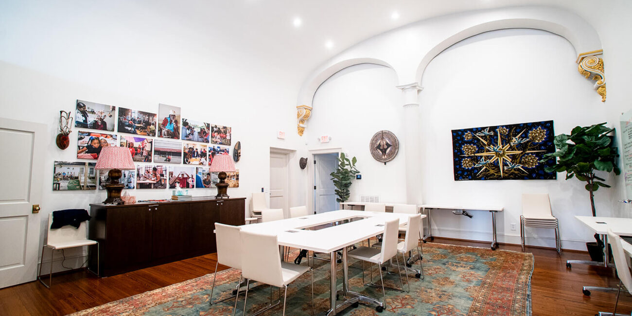 A bright modern room with white walls, arched columns, a large rug, meeting tables and chairs, wall art, and green plants.