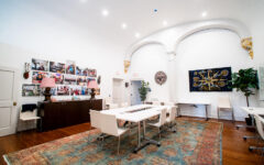 A bright modern room with white walls, arched columns, a large rug, meeting tables and chairs, wall art, and green plants.