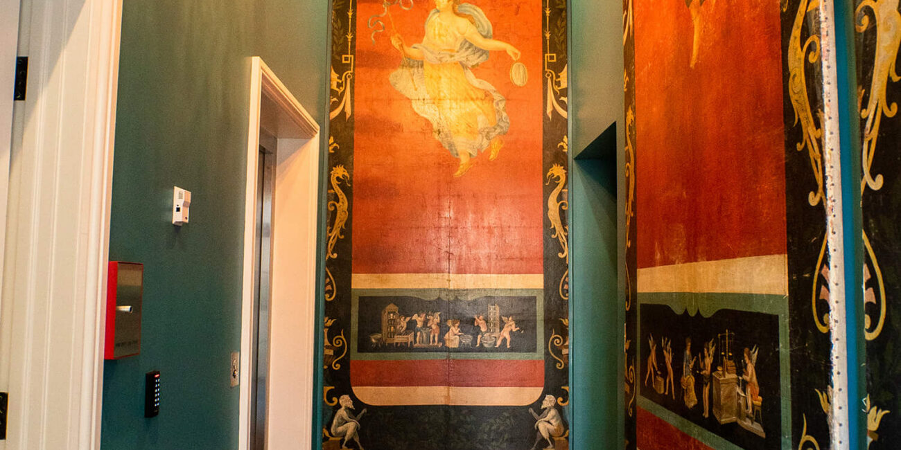 A hallway with teal walls, classical murals, a red patterned rug on wood floors, and a lit crystal chandelier above.