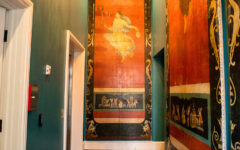 A hallway with teal walls, classical murals, a red patterned rug on wood floors, and a lit crystal chandelier above.