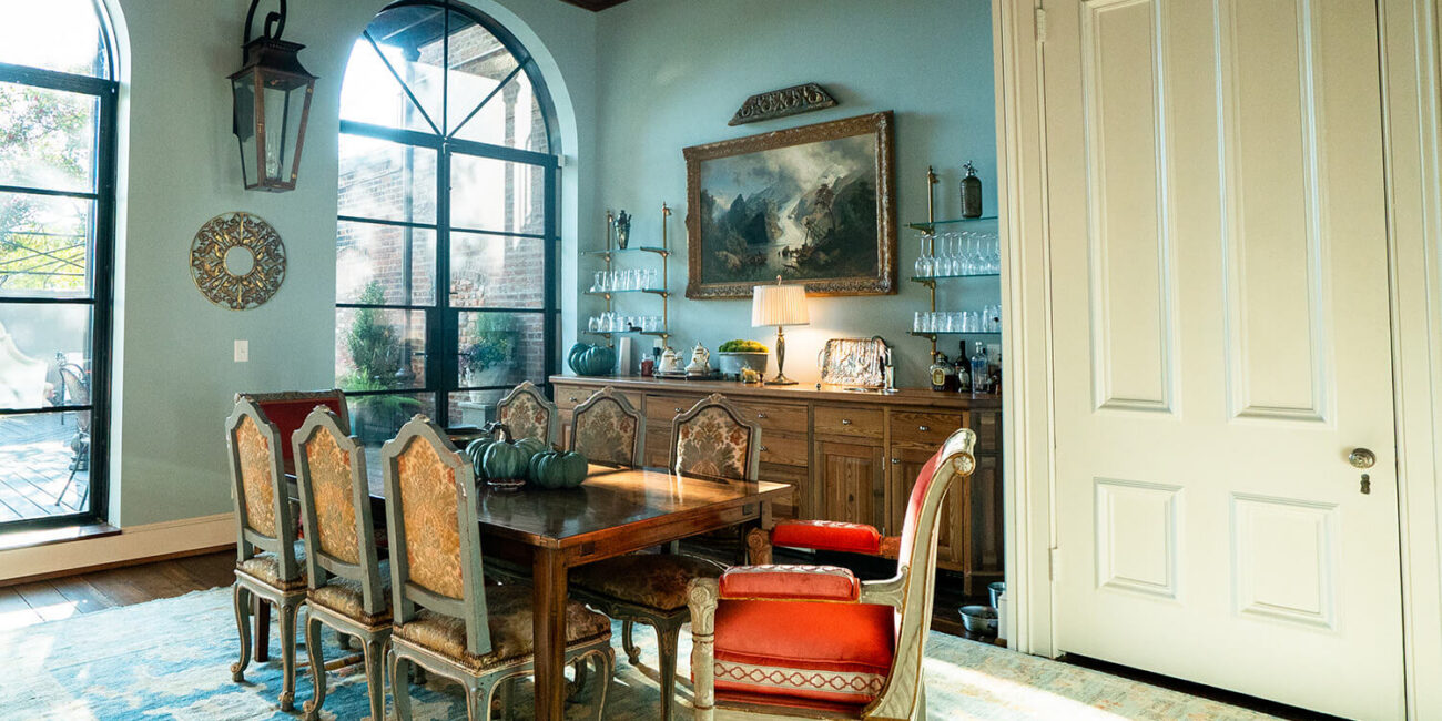 Cozy dining room featuring a wooden table, six ornate chairs (two red), sideboard with lamp, decorative plates, painting, arched windows.