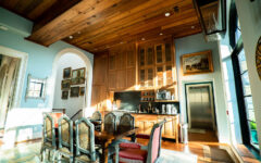 Bright kitchen and dining area with wood ceiling, tall cabinets, stylish chairs at a table, wall art, big windows, and an elevator.