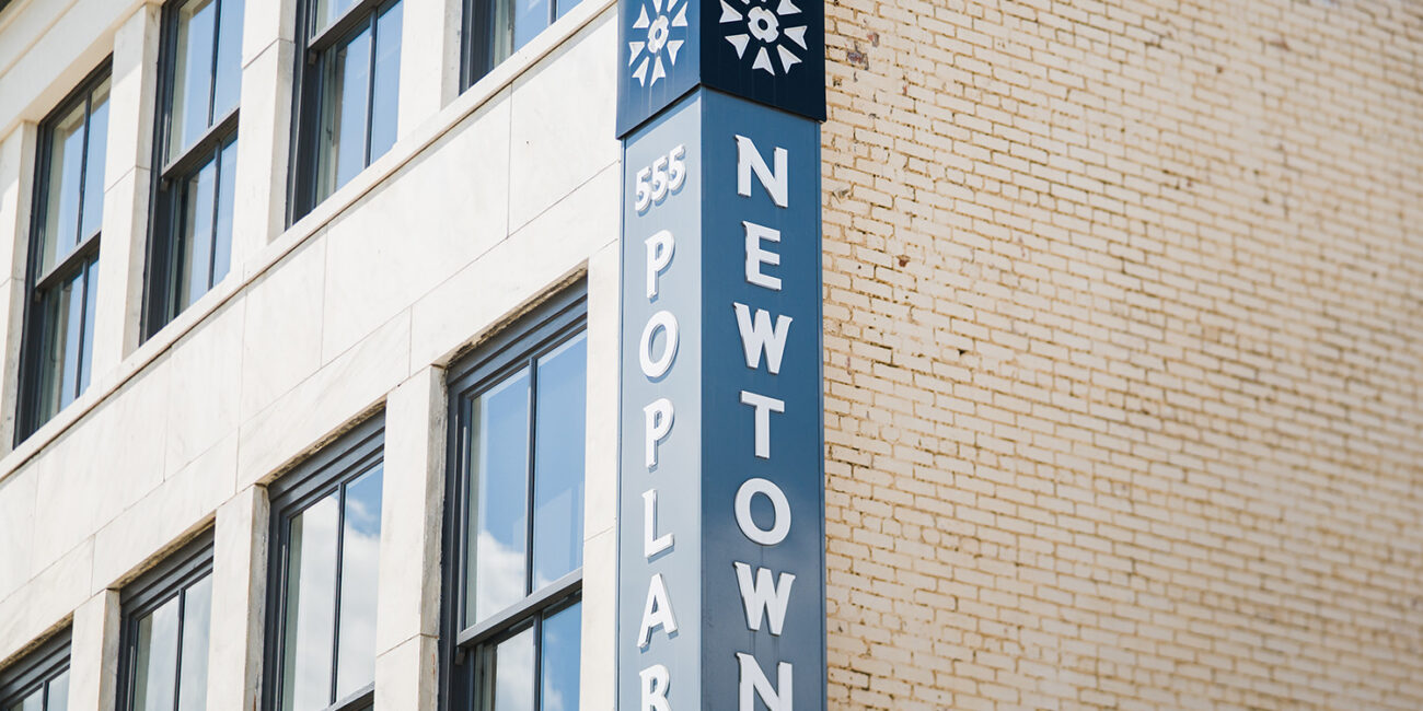 A blue and white vertical sign reading 555 Poplar NEWTOWN is mounted on the corner of a tan brick building with large windows.
