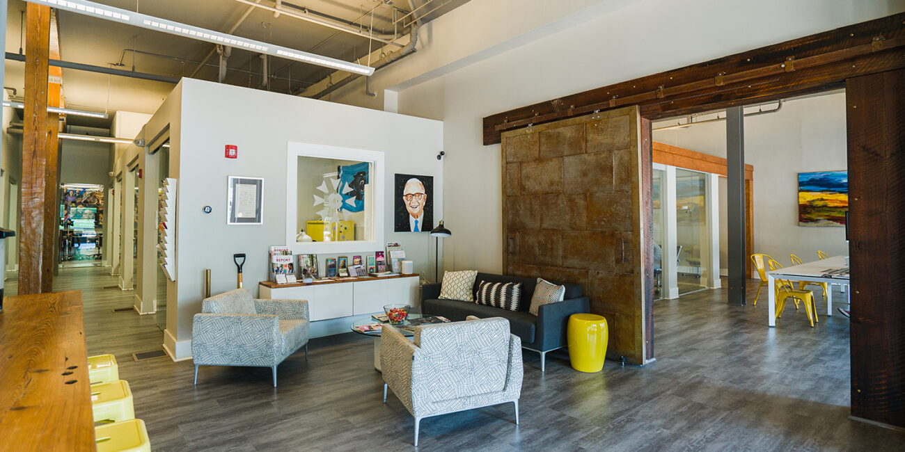 Modern office with light wood floors, gray walls, exposed ceilings, cozy seating, wall art, open workspace, and yellow chairs.