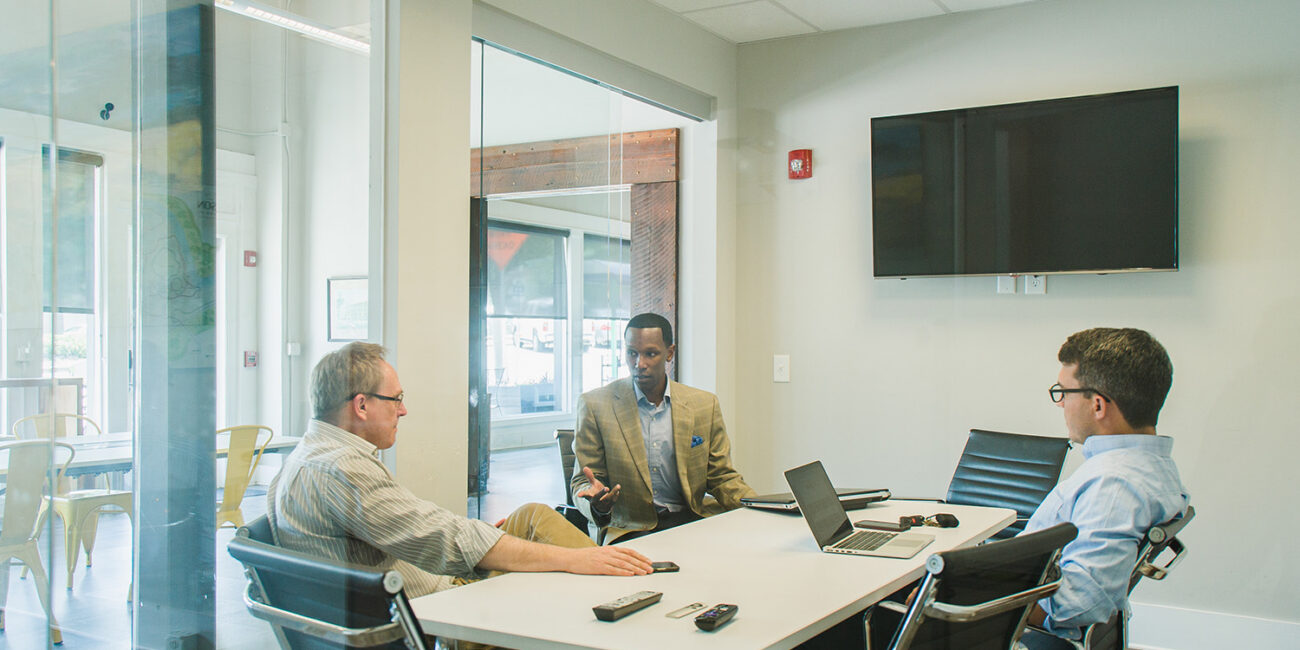 Three men sit at a conference table in a modern office, talking. Laptops and remotes are on the table; glass walls enclose the room.