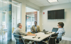 Three men sit at a conference table in a modern office, talking. Laptops and remotes are on the table; glass walls enclose the room.