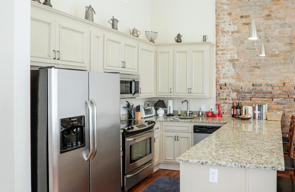 Modern kitchen with stainless steel appliances, light cabinets, granite countertops, brick accent wall, hardwood floors, and pendant lighting.
