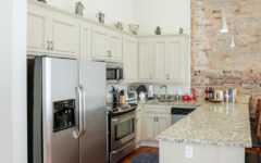 Modern kitchen with stainless steel appliances, light cabinets, granite countertops, brick accent wall, hardwood floors, and pendant lighting.