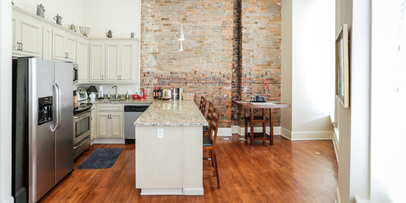 Modern kitchen with stainless steel appliances, light cabinets, granite island and barstools, brick wall, wood floors, sunlit.