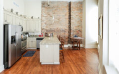 Modern kitchen with stainless steel appliances, light cabinets, granite island and barstools, brick wall, wood floors, sunlit.