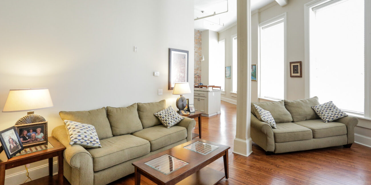 A bright living room with beige sofas, patterned pillows, wood coffee table, side tables with lamps, and sunlit windows. Decor on tables.
