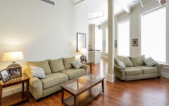 A bright living room with beige sofas, patterned pillows, wood coffee table, side tables with lamps, and sunlit windows. Decor on tables.