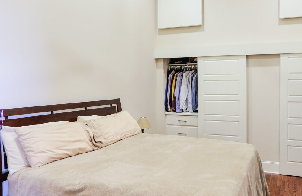 A neatly made bed with beige sheets and pillows on a wooden floor in a minimalist room, open closet and ceiling fan above.