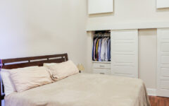 A neatly made bed with beige sheets and pillows on a wooden floor in a minimalist room, open closet and ceiling fan above.