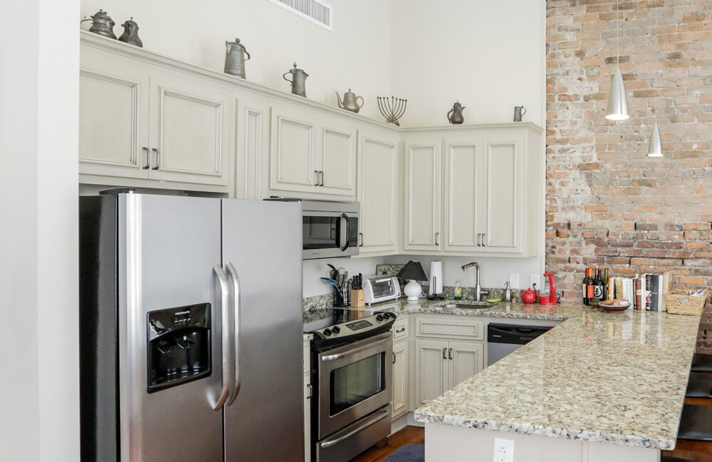 Modern kitchen featuring stainless steel appliances, light cabinets, granite counters, brick accent wall, and hardwood floors.