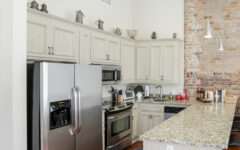 Modern kitchen featuring stainless steel appliances, light cabinets, granite counters, brick accent wall, and hardwood floors.