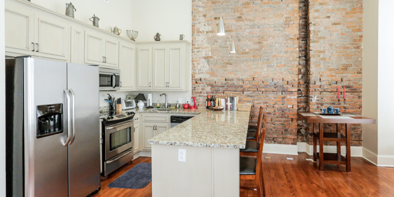Modern kitchen with stainless steel appliances, granite counters, cream cabinets, brick wall, wood island with pendant lights.