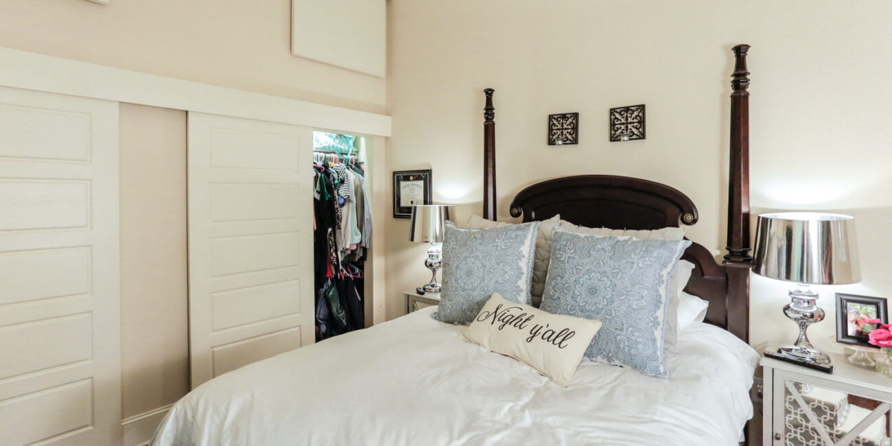 Cozy bedroom with a four-poster bed, white bedding, blue decorative pillows, and a Night y'all accent pillow. Two mirrored nightstands with lamps flank the bed. A closet with sliding doors is partially open.