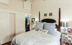 Cozy bedroom with a four-poster bed, white bedding, blue decorative pillows, and a Night y'all accent pillow. Two mirrored nightstands with lamps flank the bed. A closet with sliding doors is partially open.