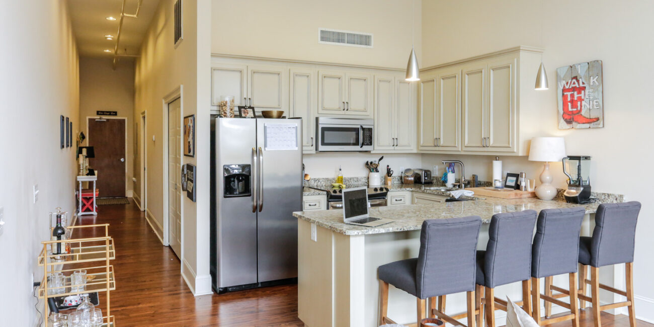 Modern kitchen with white cabinets, stainless steel appliances, granite island, three gray barstools, and a bar cart by wooden floor.