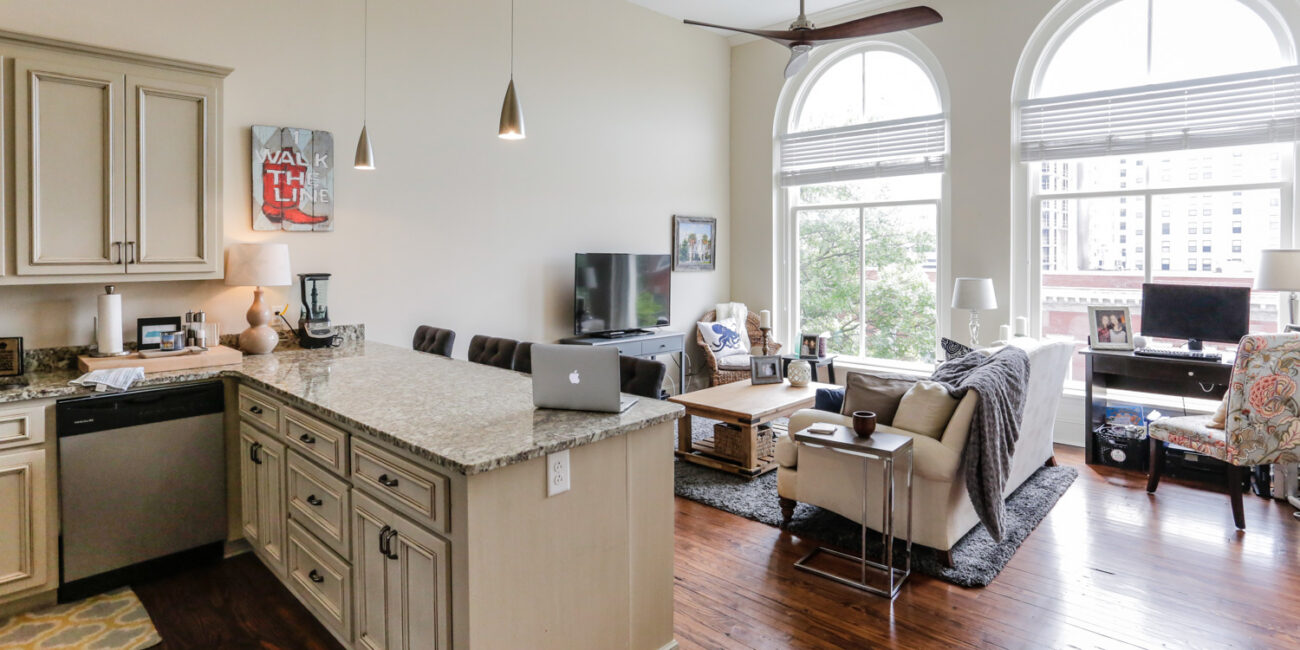 Modern open-concept living room and kitchen with arched windows, light cabinets, granite counters, hardwood floors, TV, and sofa.